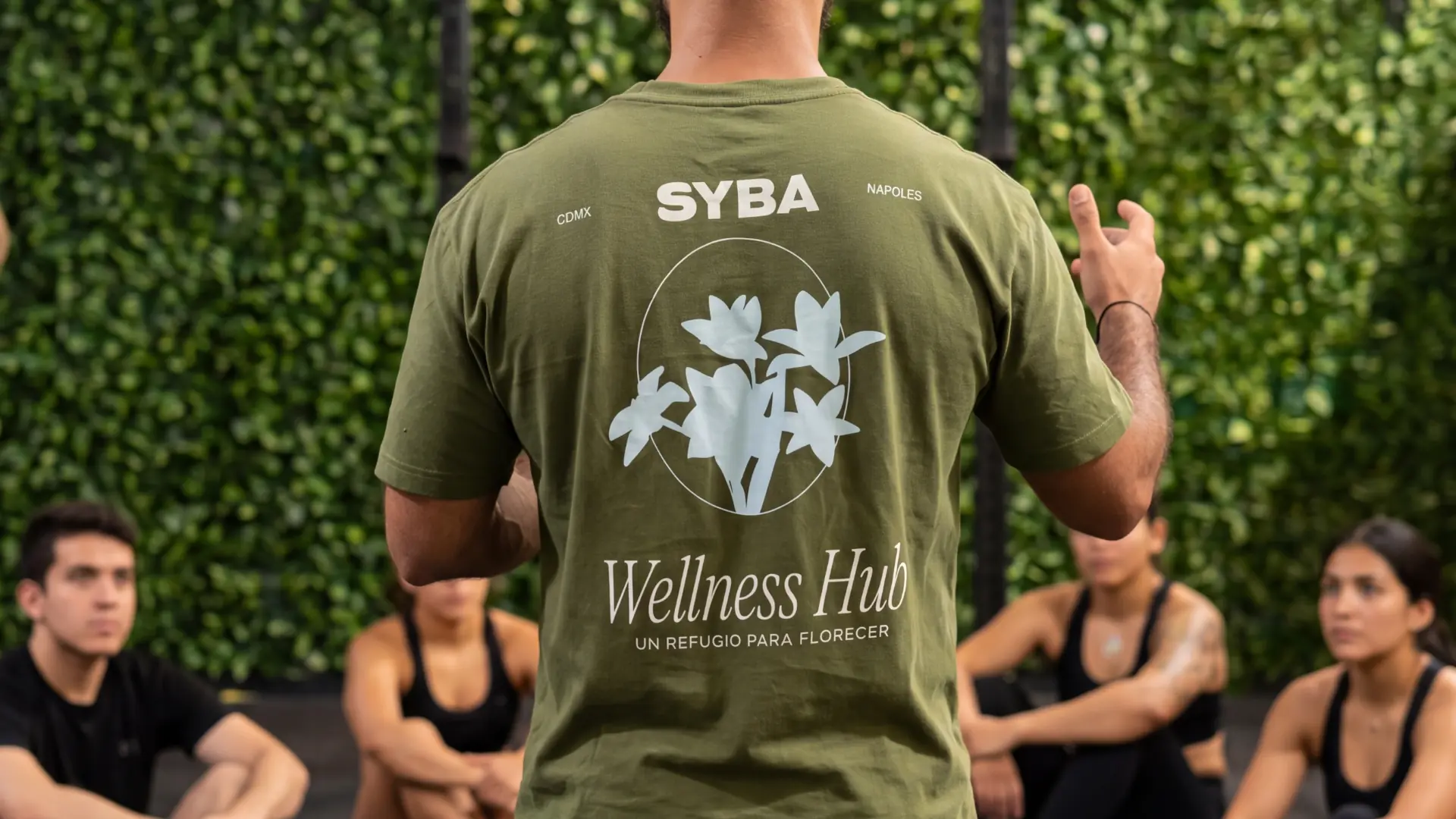 Coach con playera de SYBA 'Un refugio para florecer' dando indicaciones a su grupo frente al muro verde del estudio